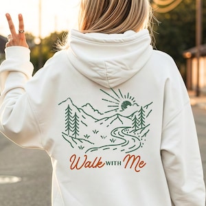Puede incluir: Sudadera con capucha blanca con un gráfico de temática natural en la espalda. El diseño presenta montañas, árboles, un río y un sol, con el texto "Walk with Me" en naranja. La sudadera tiene capucha con cordón y mangas largas.