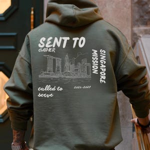 Op de afbeelding: Militairgroene hoodie met een capuchon en lange mouwen. De achterkant heeft witte tekst en een stadsgezichtafbeelding met de tekst "SENT TO GATHER, Singapore Mission, called to serve, 2025-2027."