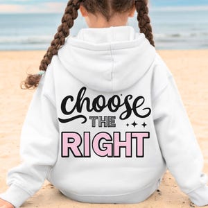 Op de afbeelding: Een witte hoodie met de tekst "choose THE RIGHT" in zwart en roze. De persoon die de hoodie draagt, zit op een zandstrand met de zee op de achtergrond.