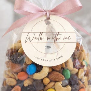 Op de afbeelding: Een doorzichtige cellofaan zak gevuld met een mix van noten, chocoladesnoepjes en gedroogd fruit. Een rond label met de tekst "Walk with me" en "One step at a time" is bevestigd met een roze lint.