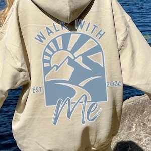 May include: A beige hoodie featuring a blue and white graphic on the back with the text "Walk With Me" and a mountain and sun design. The hoodie is worn with white sweatpants. The image is taken outdoors near a lake.