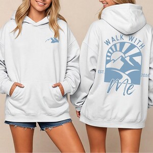 May include: White hoodie with a front pocket and a hood. The front features a small light blue mountain graphic. The back displays a larger light blue design with the text "WALK WITH Me" and a mountain and sun graphic. The hoodie is a casual style.