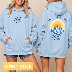May include: Light blue hoodie with a front pocket and a hood. The front features the word "Me" in cursive with "Moses 6:34" below it. The back displays "Walk With Me" around a mountain and sun graphic, with the same text.