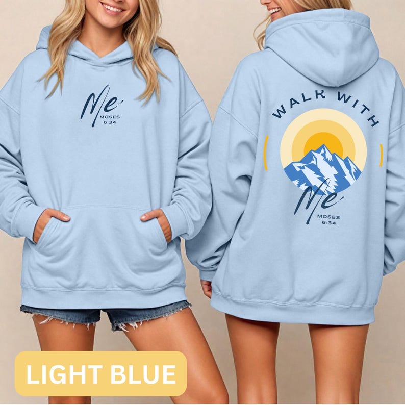 May include: Light blue hoodie with a front pocket and a hood. The front features the word "Me" in cursive with "Moses 6:34" below it. The back displays "Walk With Me" around a mountain and sun graphic, with the same text.