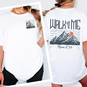 May include: White t-shirt with a mountain and sun graphic on the front pocket and back. The back graphic reads "WALK with ME" above a mountain scene and "Moses 6:34" below. The design features gray, red, and white colors.