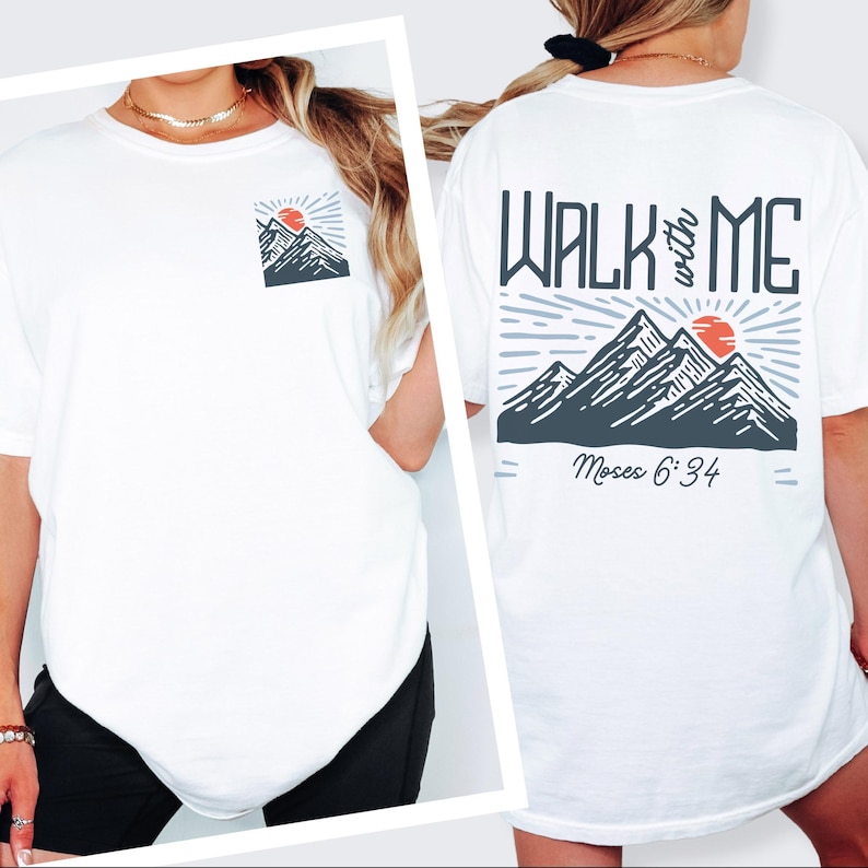May include: White t-shirt with a mountain and sun graphic on the front pocket and back. The back graphic reads "WALK with ME" above a mountain scene and "Moses 6:34" below. The design features gray, red, and white colors.