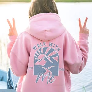 May include: A light pink hoodie with the text "WALK WITH ME" and a mountain graphic in blue and white. The hoodie has a drawstring hood and long sleeves. The year "2026" is printed below the graphic.