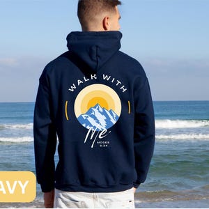 May include: Navy blue hoodie with a graphic on the back featuring a mountain scene inside a circle with the words "WALK WITH ME" and the Bible verse "Moses 6:34". The hoodie is shown against a beach backdrop.