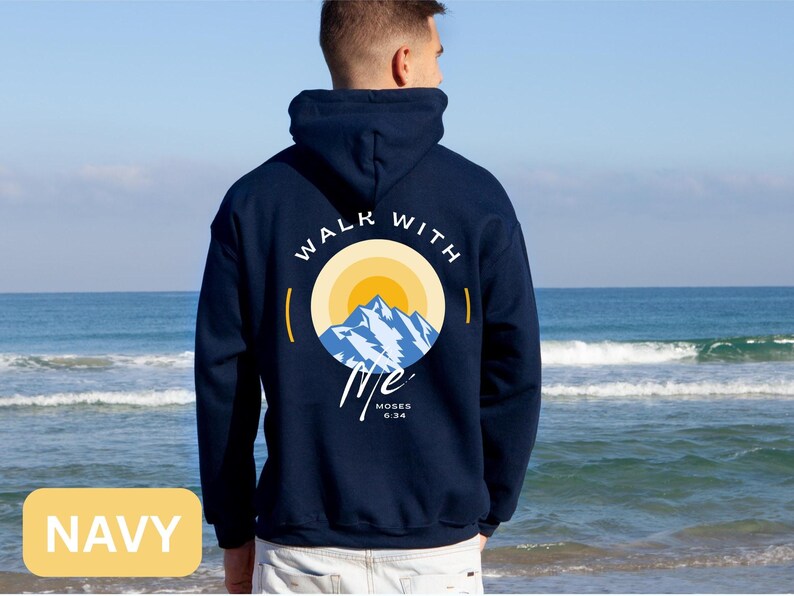 May include: Navy blue hoodie with a graphic on the back featuring a mountain scene inside a circle with the words "WALK WITH ME" and the Bible verse "Moses 6:34". The hoodie is shown against a beach backdrop.