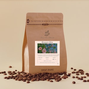 Könnte beinhalten: Ein brauner Papiersack mit Kaffeebohnen und dem Text "Blossom Bite" auf einem Etikett. Das Etikett enthält ein Foto von rosa Blumen. Der Beutel ist von verstreuten Kaffeebohnen umgeben. Der Beutel ist 340g oder 12oz.