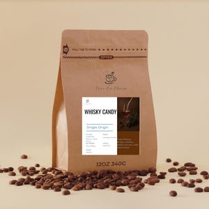 Op de afbeelding: Een bruine papieren zak met "Whisky Candy" koffiebonen met een wit etiket en een kleine afbeelding van een drankje. De zak is omgeven door verspreide koffiebonen. De zak is gelabeld "12OZ 340G".