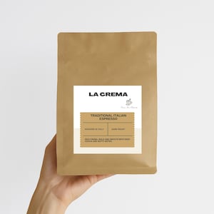 May include: A brown paper bag of La Crema Traditional Italian Espresso coffee. The bag has a white label with the brand name and coffee details. The label states the coffee is roasted in Italy and is a dark roast. The bag is held by a person.