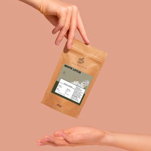 Könnte beinhalten: Ein brauner Papiertüte mit White Lotus Tee wird zwischen zwei Händen vor einem pfirsichfarbenen Hintergrund gehalten. Die Tüte hat ein Etikett mit Text und einem floralen Design. Die Tüte ist mit "3OZ" beschriftet. Der Markenname "Pour On Charm" ist oben aufgedruckt.