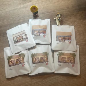 Könnte beinhalten: Weiße Kaffeebohnenbeutel mit Illustrationen eines Cafés. Die Beutel sind auf einer Holzoberfläche angeordnet, zusammen mit kleinen Figuren einer Sonnenblume und einer Katze mit Sonnenbrille. Auf den Beuteln steht das Wort "coffee".