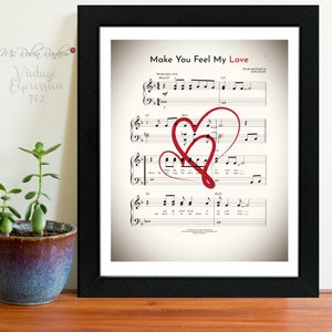May include: A framed print of sheet music for the song "Make You Feel My Love" by Bob Dylan. The sheet music features two red hearts drawn over the notes.