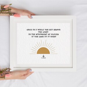 Peut inclure: Une impression encadrée avec un design minimaliste représentant un soleil jaune avec des rayons et une silhouette noire d'une personne assise en position de yoga. Le texte dit "Once in a while you get shown the light. In the strangest of places if you look at it right."