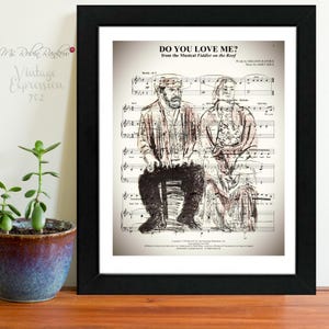 May include: Black framed print featuring sheet music with a sketch of two people. The text "DO YOU LOVE ME?" is at the top, with the musical title and credits. The print is on a wooden surface with a potted plant.