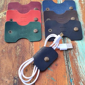Puede incluir: Una colección de organizadores de cables de cuero en varios colores: burdeos, rojo, verde, azul, marrón y negro. Cada organizador tiene un cierre de botón a presión y sujeta un cable USB blanco. Se muestran sobre una superficie de madera.