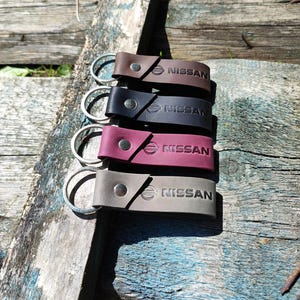 Op de afbeelding: Een stapel van vier leren sleutelhangers in bruin, zwart, mauve en grijs. Elke sleutelhanger heeft een zilveren ring en het woord "NISSAN" in reliëf met het Nissan-logo. De sleutelhangers zijn vastgezet met een drukknoopsluiting.