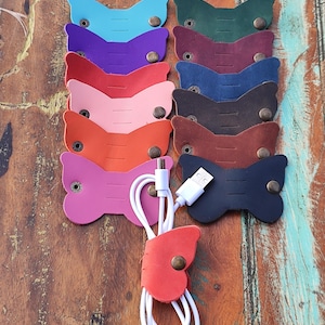 Puede incluir: Una colección de organizadores de cables de cuero de colores en varios tonos, incluyendo azul, morado, rosa y marrón. Cada organizador tiene forma de mariposa y se asegura con un broche metálico. Un cable USB blanco está cuidadosamente organizado con un organizador rojo.