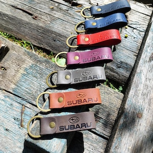 Könnte beinhalten: Sieben Leder-Schlüsselanhänger in verschiedenen Farben, darunter Blau, Rot und Braun, liegen auf einer verwitterten Holzoberfläche. Jeder Schlüsselanhänger trägt das Subaru-Logo und -Schriftzug, mit einem Messingfarbenen Ring und Knopfverschluss.