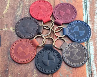May include: A selection of leather keyrings in various colours, featuring the BMW logo. The keyrings are round with stitched edges and attached to metal key rings. Colours include red, burgundy, black, blue, brown, and tan.