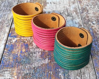 Può includere: Tre bracciali da polso in pelle dai colori vivaci: giallo, rosa e verde acqua. Ogni bracciale è composto da più strisce orizzontali di pelle e ha una chiusura a scatto. Esposti su una superficie di legno invecchiato.