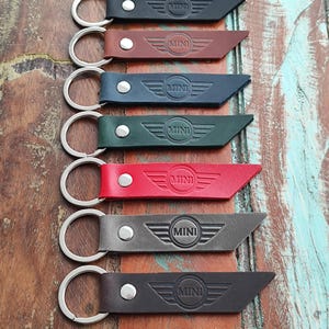 May include: Seven leather keyrings in a range of colours, each embossed with the "MINI" logo and wings. The keyrings have silver rings and are displayed on a rustic wooden surface.