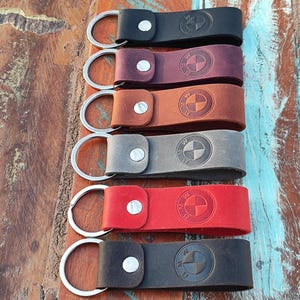 Op de afbeelding: Een verzameling van zes leren sleutelhangers in verschillende kleuren: zwart, bordeauxrood, bruin, grijs, rood en donkergrijs. Elke sleutelhanger heeft een zilveren metalen ring en een knoopsluiting, met het BMW-logo in reliëf op het leer.