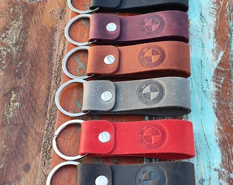 May include: Six leather keyrings in black, burgundy, brown, grey, red, and dark grey. Each keyring has a silver metal ring and a button clasp, with the BMW logo embossed on the leather.