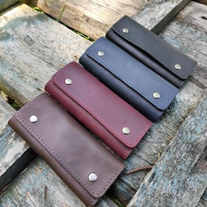 May include: Several leather key holders in shades of brown, burgundy, navy, and black. Each has a rectangular shape, a snap closure, and visible stitching. The key holders are displayed on a weathered wooden surface.