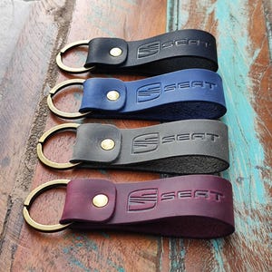 Könnte beinhalten: Vier Leder-Schlüsselanhänger in Schwarz, Blau, Grau und Burgunderrot. Jeder Schlüsselanhänger hat einen goldfarbenen Ring und einen Druckknopfverschluss. Das Wort "SEAT" ist auf jedem Schlüsselanhänger geprägt.