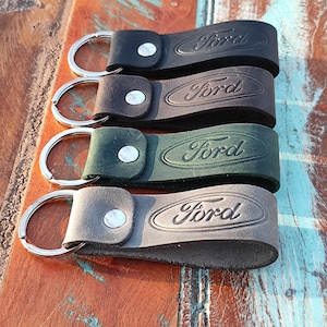 Puede incluir: Cuatro llaveros de cuero en negro, marrón, verde y gris. Cada llavero tiene un anillo plateado y un cierre de botón a presión. La palabra "Ford" está grabada en relieve en cada llavero.