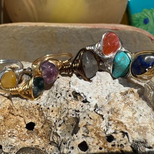 May include: A collection of gemstone rings with wire wrapping. The rings feature various stones in shades of yellow, green, purple, gray, red, turquoise, and blue. The wire is silver and gold-toned, creating a handcrafted look.