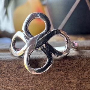 May include: A polished silver ring featuring a central openwork design composed of four interconnected loops arranged in a symmetrical clover-like pattern. The metal surface is smooth and reflective, showcasing a continuous, fluid metallic band.