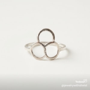 May include: A silver ring with a unique openwork design. The ring features three interconnected circles, creating a modern and geometric aesthetic. The ring is set against a white background.