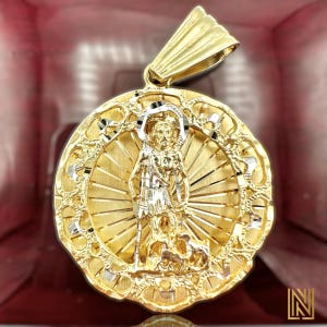 May include: Gold-tone pendant featuring a religious figure with a halo, set against a radiant background. The pendant has intricate detailing and a textured finish. The bail is a solid gold color. The pendant is likely made of gold or gold-plated metal.