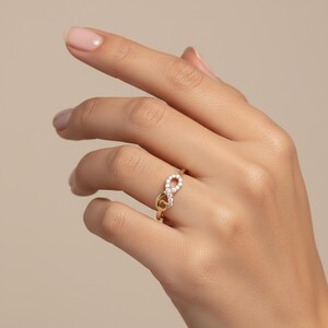 Pu&ograve; includere: Un anello dorato con un design a simbolo dell'infinito. Il simbolo dell'infinito &egrave; incastonato con piccole gemme trasparenti. L'anello &egrave; indossato su un dito, mostrando il suo aspetto delicato ed elegante. Il design dell'anello &egrave; un simbolo di eternit&agrave; e amore.