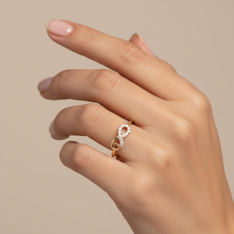 Pu&ograve; includere: Un anello dorato con un design a simbolo dell'infinito. Il simbolo dell'infinito &egrave; incastonato con piccole gemme trasparenti. L'anello &egrave; indossato su un dito, mostrando il suo aspetto delicato ed elegante. Il design dell'anello &egrave; un simbolo di eternit&agrave; e amore.