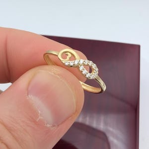 Pu&ograve; includere: Un anello dorato a forma di infinito con una fila di piccole pietre preziose trasparenti incastonate nel simbolo dell'infinito. L'anello &egrave; tenuto dalle dita di una persona, con uno sfondo rosso scuro.