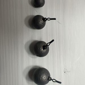 May include: Four gray fishing weights with attached black clips are arranged vertically against a white background. The weights are spherical with a textured surface. The clips are made of wire and are looped.