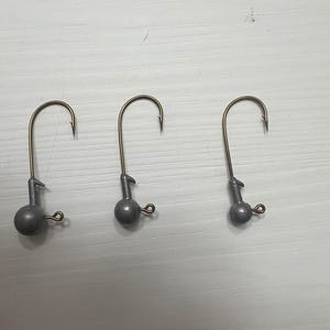 May include: Three gray jig heads with gold-colored hooks. Each jig head has a round weight and a small wire loop. The hooks are curved and pointed, designed for fishing. The items are arranged in a row.