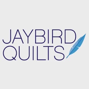 May include: The logo for Jaybird Quilts features the text "JAYBIRD QUILTS" in a dark blue sans-serif font. A stylized blue feather is positioned to the right of the text against a light gray background.