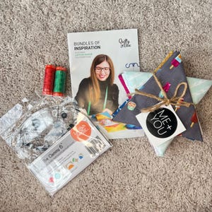 May include: A quilting kit containing a Quilty Box booklet, two spools of red and green thread, a plastic Clammy 6 ruler in packaging, and a stack of folded fabric squares tied with twine and a circular tag reading ME + YOU.