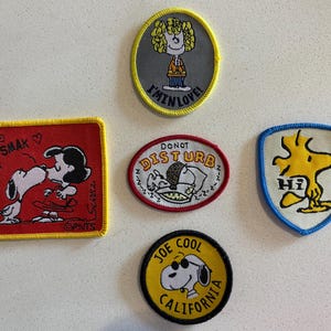 May include: A collection of embroidered patches featuring Peanuts characters. Includes a red square patch with Snoopy kissing Lucy, a yellow patch with Woodstock, and a yellow patch with Snoopy wearing sunglasses. Other patches include the text "I'M IN LOVE!" and "DO NOT DISTURB".