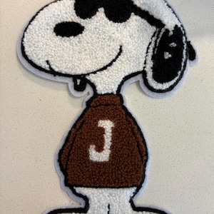 May include: A Snoopy patch with a white body, black sunglasses, and a brown sweater with a white "J" on it. The patch has a textured, tufted appearance and a black outline. The cartoon character is smiling.
