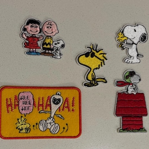 May include: A collection of embroidered patches featuring characters from the Peanuts comic strip. The patches include Snoopy, Woodstock, Charlie Brown, Lucy, and a patch with the text "HA HA HA!". The patches are colorful and have a cartoon style.