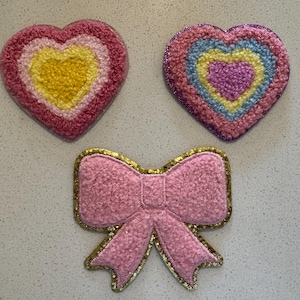 Könnte beinhalten: Drei dekorative Aufnäher: zwei herzförmige und einer in Form einer Schleife. Die Herzen haben Farbschichten aus Rosa, Gelb und Blau. Die Schleife ist rosa mit einem goldenen Glitzerumriss. Alle Aufnäher haben ein strukturiertes, flauschiges Aussehen.