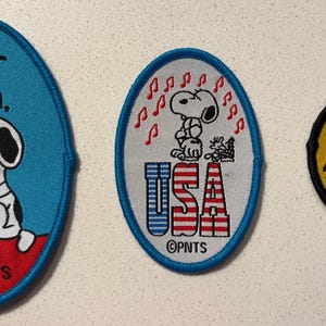 Könnte beinhalten: Drei gestickte Snoopy-Aufnäher. Ein ovales Patch mit Snoopy und dem Text "Dear Mom". Ein weiteres ovales Patch mit Snoopy und "USA". Das dritte Patch ist rund und trägt die Aufschrift "Joe Cool California."