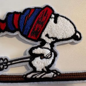 May include: Embroidered patch featuring Snoopy skiing, wearing a red, white, and blue striped hat. Woodstock is also present. The patch has a white background and a brown ski.